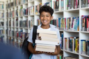 A middle school student, Marcus, looks at the viewer smiling. He is standing in a library holding books.